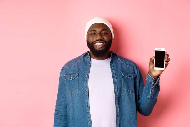 Handsome hipster guy in beanie and denim shirt smiling, showing mobile phone screen with happy face, introduce application, standing over pink background - Photo, Image