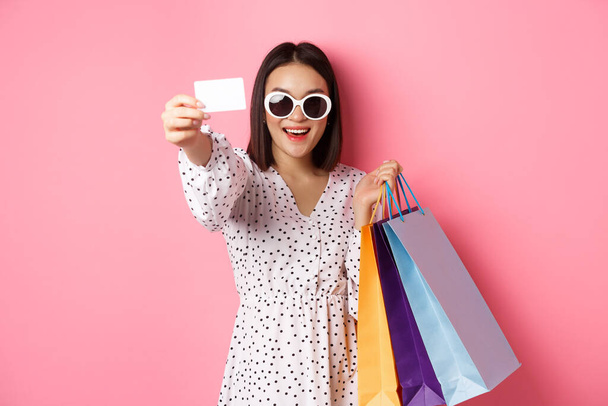 Hermosa mujer asiática en gafas de sol ir de compras, sosteniendo bolsas y mostrando la tarjeta de crédito, de pie sobre fondo rosa - Foto, imagen