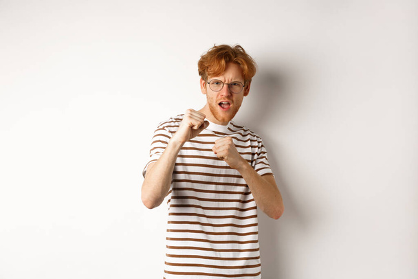Funny young man with red hair raising fists for fight, shadow boxing and looking serious at camera, standing over white background - Photo, Image