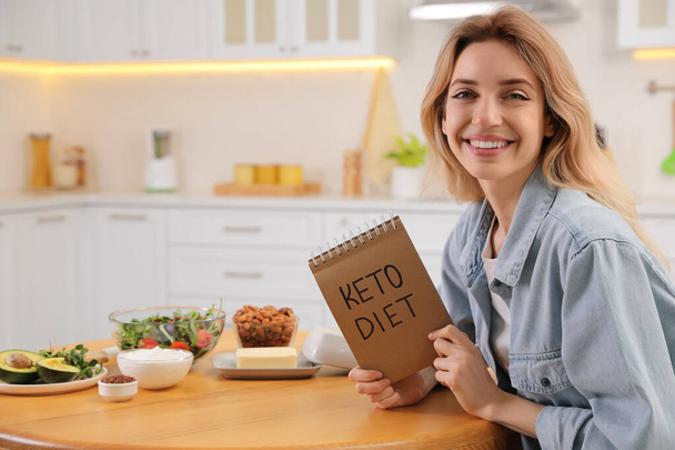 Frau hält Notizbuch mit Satz Keto Diet und verschiedenen Produkten am Holztisch in der Küche, Platz für Text - Foto, Bild