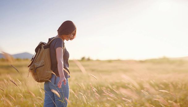 Trendy young woman with a backpack in a field at sunset. Freedom and nature concept. Soft focus - Фото, зображення