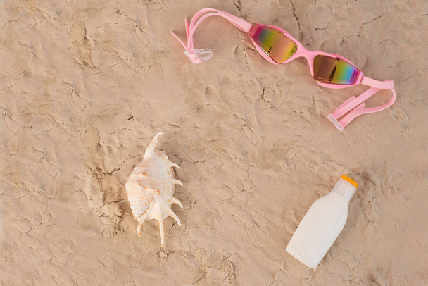 Top view of seashell swimming goggles and sunscreen lotion on sand. - Foto, afbeelding