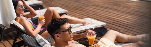Mann mit Sonnenbrille hält Orangensaft neben verschwommener Freundin auf Liegestuhl, Transparent  - Foto, Bild