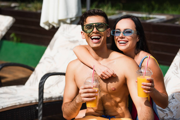 Happy woman with orange juice embracing boyfriend in sunglasses on deck chair  - Foto, afbeelding