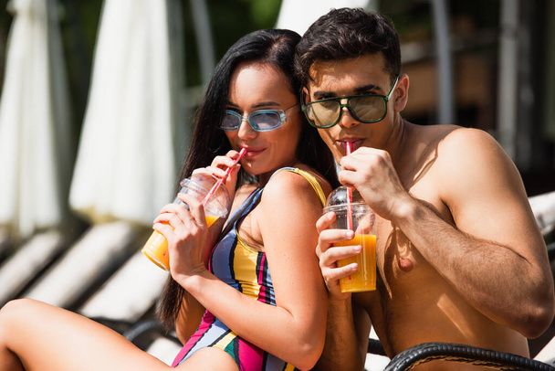 Pareja joven en gafas de sol bebiendo jugo de naranja en el resort  - Foto, Imagen
