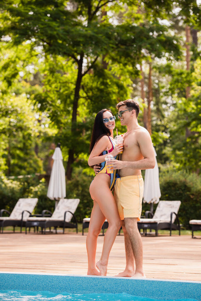 Shirtless man hugging smiling girlfriend with orange juice near poolside  - Фото, изображение