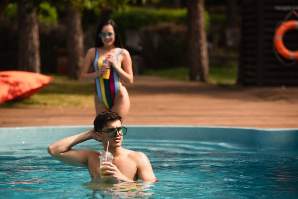 Young man in sunglasses holding orange juice near girlfriend on blurred background in swimming pool  - Foto, imagen