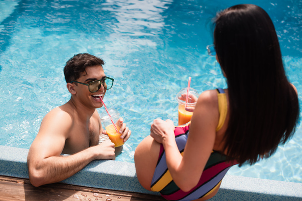 Uomo sorridente che beve succo d'arancia vicino alla donna sfocata in piscina  - Foto, immagini