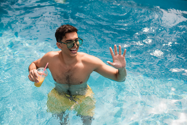 Vista de ángulo alto del hombre sonriente con jugo de naranja agitando la mano en la piscina  - Foto, imagen