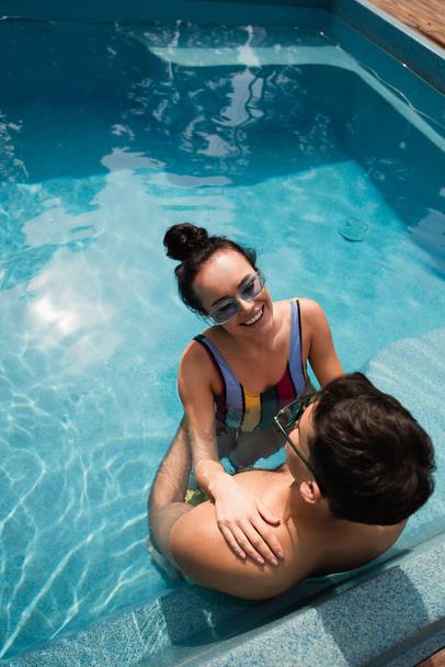 Overhead view of smiling woman resting with boyfriend in sunglasses in swimming pool  - Photo, Image