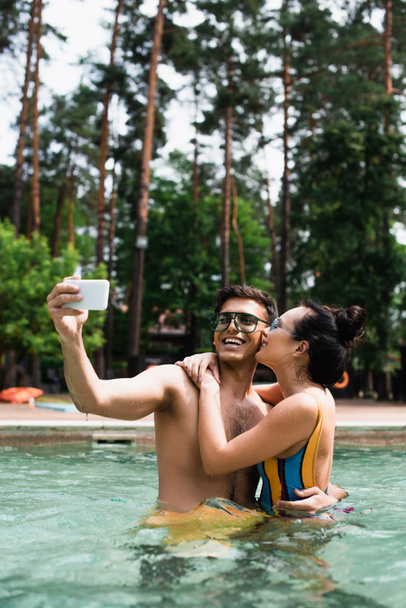 Pretty woman kissing and embracing boyfriend taking selfie in swimming pool  - Photo, Image