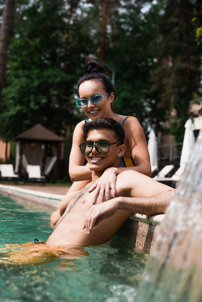 Mujer sonriente relajándose con su novio en la piscina cerca de la fuente borrosa  - Foto, imagen