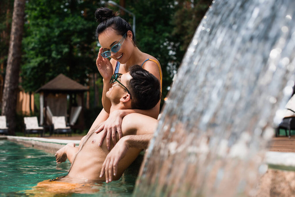 Woman holding sunglasses and embracing boyfriend near fountain in swimming pool  - Photo, Image