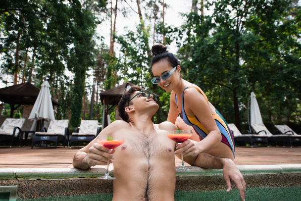 Usmívající se pár ve slunečních brýlích drží koktejly u bazénu  - Fotografie, Obrázek