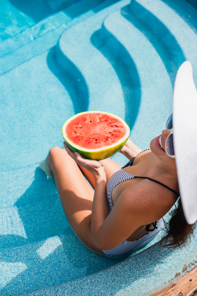 Pohled shora na usmívající se ženu ve slunečních brýlích držící meloun v bazénu  - Fotografie, Obrázek
