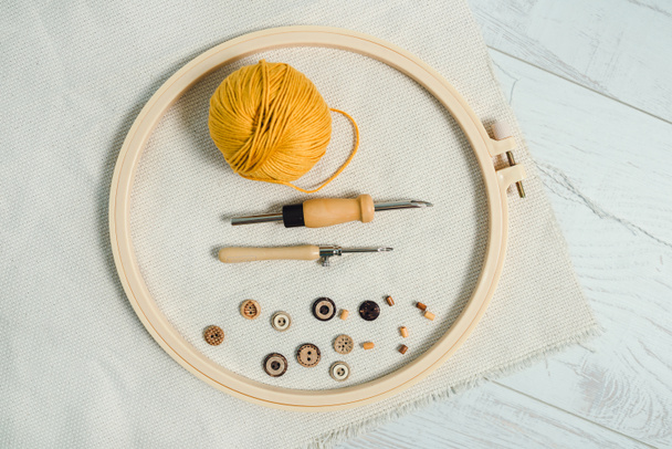 Embriodery aro colocado alrededor de dos agujas de perforación, bola de hilo amarillo, botones de madera y pieza de tela de lino en el suelo de madera blanca. Punch aguja trabajo hobby creativo. - Foto, imagen