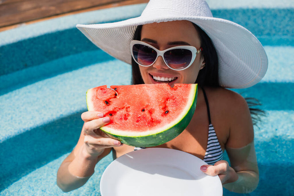 Happy woman holding piece of watermelon and plate in pool during weekend  - Foto, Imagem