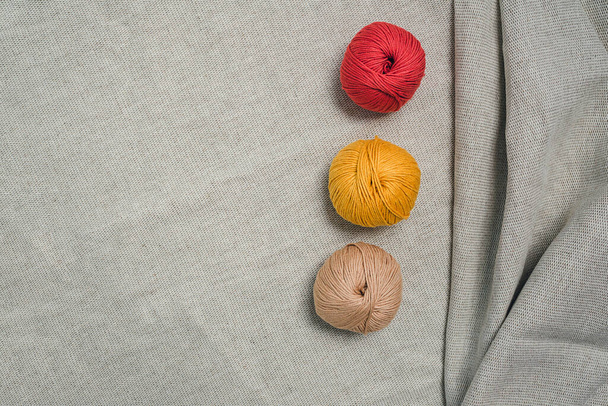 Thee balls of yarn on grey linen fabric with folds on the edge. Red yellow and beige colors. Creative hobby concept. - Photo, Image