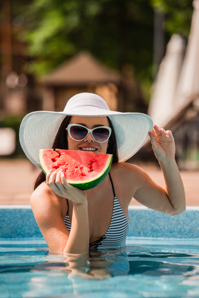 Karpuz ve güneş şapkasını havuzda tutarken gülümseyen genç bir kadın.  - Fotoğraf, Görsel