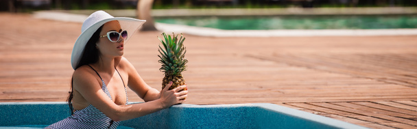 Brunette woman in sun hat holding pineapple in swimming pool on resort, banner  - Photo, image