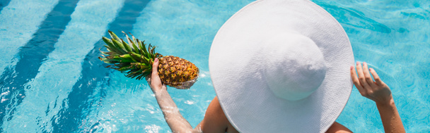 白い太陽の帽子の女性のオーバーヘッドビュースイミングプール、バナーでパイナップルを保持  - 写真・画像