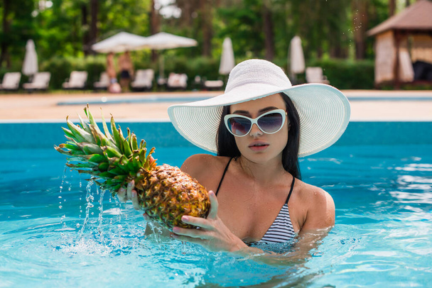 Молодая женщина держит ананас с капельками воды в бассейне  - Фото, изображение