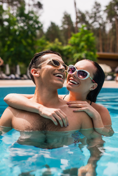 Donna sorridente in occhiali da sole che abbraccia allegro fidanzato in piscina  - Foto, immagini