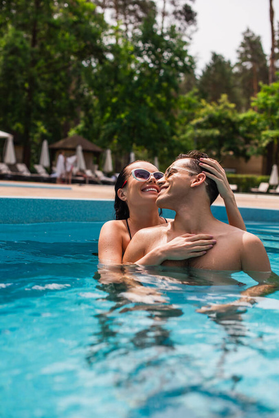 Šťastná brunetka žena v slunečních brýlích objímání mladý přítel v bazénu  - Fotografie, Obrázek