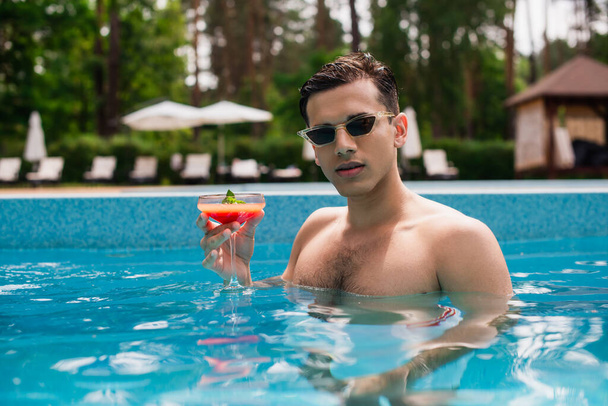 Молодой человек с коктейлем, стоящий в бассейне летом  - Фото, изображение
