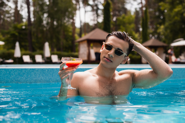 Young man in sunglasses adjusting hair and holding cocktail in swimming pool  - Zdjęcie, obraz