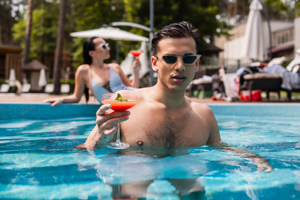 man met nat haar holding cocktail in zwembad in de buurt wazig vriendin  - Foto, afbeelding
