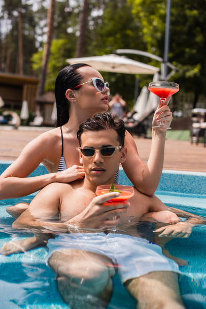 Young couple in sunglasses holding cocktails in swimming pool  - Foto, afbeelding