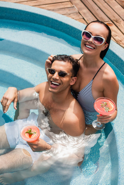 Vista de ángulo alto de pareja positiva con cócteles relajantes en la piscina durante el fin de semana  - Foto, Imagen