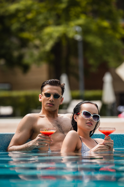 Niveau de surface du jeune couple en lunettes de soleil tenant des cocktails dans la piscine  - Photo, image