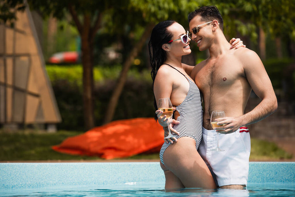 Cheerful couple hugging each other and holding glasses of wine in swimming pool  - Foto, immagini