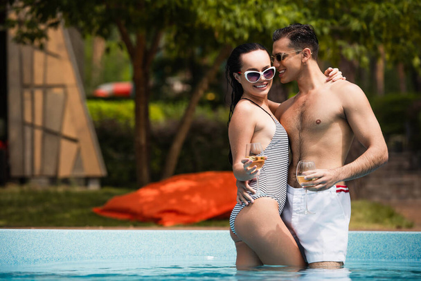 Sorrindo homem em óculos de sol e calções de banho segurando copo de vinho e abraçando namorada na piscina  - Foto, Imagem