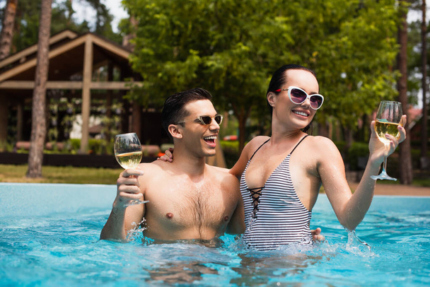 Woman in striped swimsuit holding wine and embracing man in pool  - Photo, Image