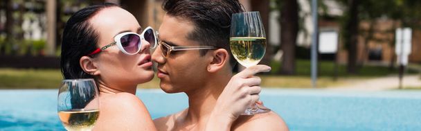 Hombre besando novia en gafas de sol sosteniendo copa de vino en la piscina sobre fondo borroso, pancarta  - Foto, Imagen