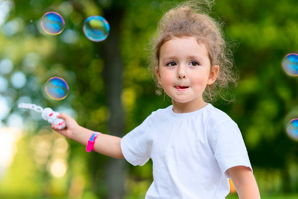 Portrait of beautiful little girl kid is having fun outdoors in a summer sunny park, happy child is blowing soap bubbles outdoors - Photo, image