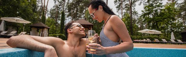 Uśmiechnięta kobieta trzymająca kieliszek wina przy basenie i chłopaka na resorcie, sztandar  - Zdjęcie, obraz
