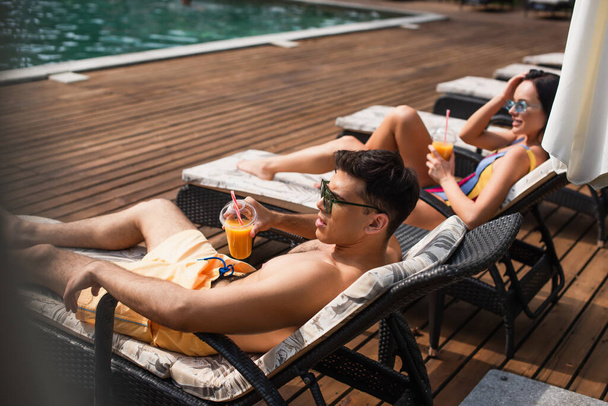 Vista lateral del hombre sosteniendo jugo de naranja en la silla de cubierta cerca de la mujer borrosa en el resort  - Foto, Imagen