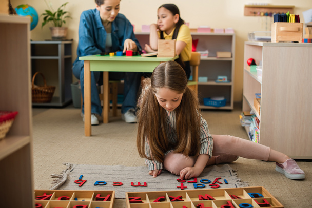 menina no chão brincando com letras de madeira perto de menina asiática e professora no fundo borrado - Foto, Imagem