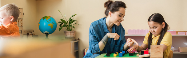 montessori school teacher pointing at multicolored blocks while playing with asian girl, banner - Foto, afbeelding