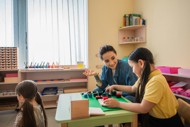 улыбающаяся учительница жестикулирует, играя в разноцветные кубики с азиатской девушкой - Фото, изображение