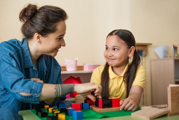 счастливая девочка и молодая учительница смотрят друг на друга, играя с деревянными кубиками - Фото, изображение