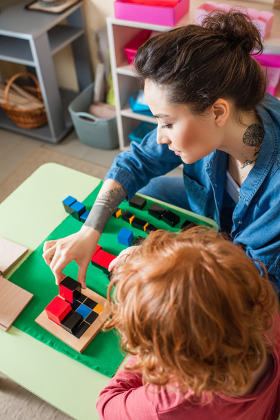 hoge hoek uitzicht op jonge leraar en roodharige kind spelen met kleurrijke kubussen in Montessori school - Foto, afbeelding