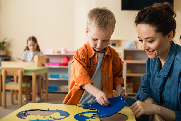 jongen combineren aarde kaart puzzel in de buurt glimlachende leraar en meisje op wazig achtergrond - Foto, afbeelding