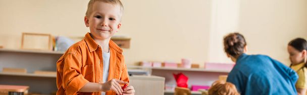 весёлый мальчик улыбается в камеру рядом с размытым учителем и детьми в школе Монтессори, баннер - Фото, изображение