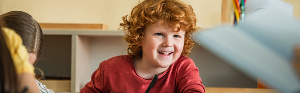 κοκκινομάλλης αγόρι χαμογελά κοντά κορίτσι στο σχολείο Montessori σε θολή πρώτο πλάνο, λάβαρο - Φωτογραφία, εικόνα
