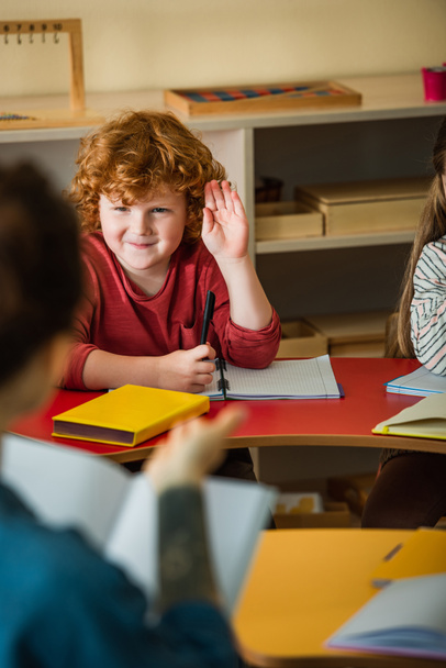 redhead boy raising hand near blurred teacher in montessori school - Foto, afbeelding